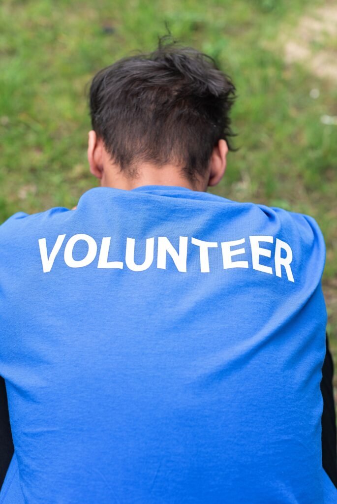 Man wearing a blue volunteer shirt sits on grass, photographed from behind.